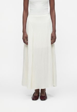 Person med hvit ermeløs topp, lang hvit flytende skjørt, og skinnende mørkerøde sko med firkantet tå, står foran en hvit bakgrunn.