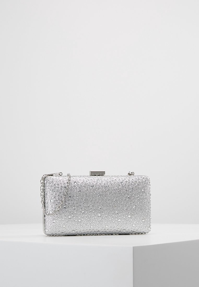 Pochette rectangulaire argentée avec une surface texturée de petits détails en forme de perles et une fine chaîne, posée sur une plateforme blanche.