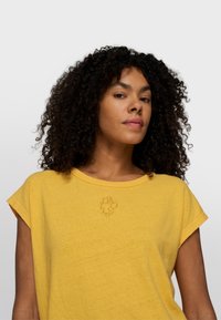 Mujer con cabello rizado que lleva una camiseta amarilla de manga corta con un pequeño logo bordado en el pecho sobre un fondo liso.