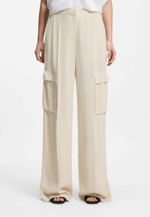 Pantaloni cargo beige a gamba larga con grandi tasche con pattina su entrambe le cosce, indossati con sandali neri open-toe e una maglietta bianca.