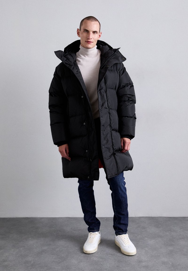 Homme portant un long manteau en doudoune noir sur un pull léger à col roulé, un jean foncé et des baskets blanches, adossé à un mur gris uni.