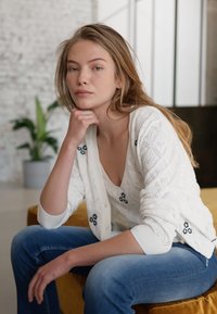 Witte gebreide cardigan met ruitpatronen en blauwe bloemaccenten, gedragen over een lichte top en gecombineerd met vervaagde blauwe jeans.