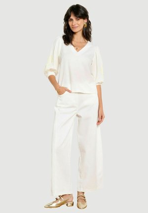 Femme aux cheveux foncés portant un chemisier blanc à col en V avec des manches bouffantes, un pantalon blanc large, des chaussures dorées et des bijoux en or, debout devant un fond clair.