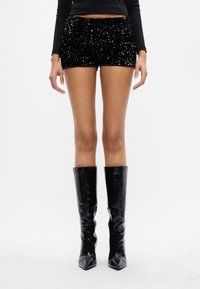 Persona vistiendo pantalones cortos negros con lentejuelas, una blusa negra de manga larga y botas negras brillantes hasta la rodilla, de pie frente a un fondo blanco.