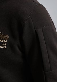 Zwarte sweater met een gestructureerde afwerking, voorzien van opvallende stiksels en een gedrukt logo in goud en wit aan de voorkant.