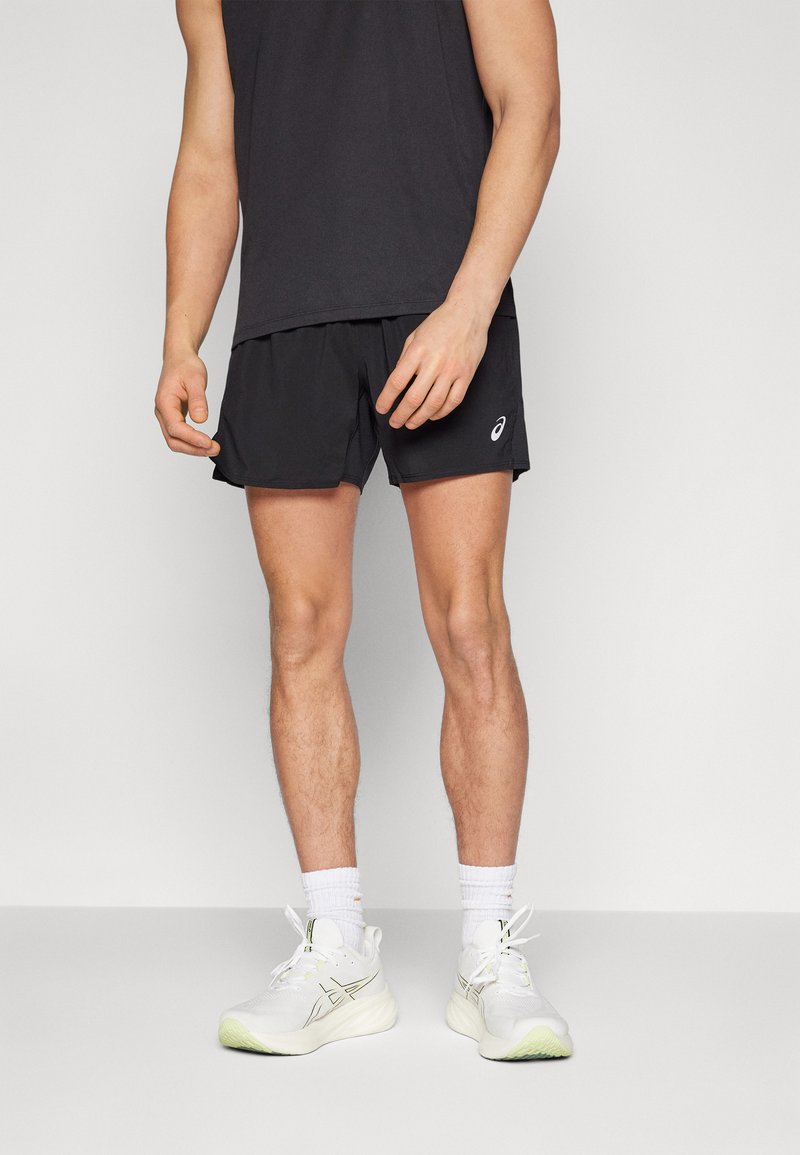 Schwarze Sportshorts mit einem elastischen Bund, kombiniert mit einem schwarzen ärmellosen Oberteil und weißen Sportschuhen mit grünen Akzenten.