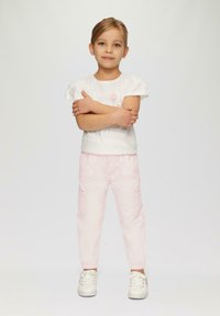 T-shirt blanc à manches courtes avec un design floral, associé à un pantalon cargo rose clair à taille élastique. Modèle portant des baskets blanches, les bras croisés.