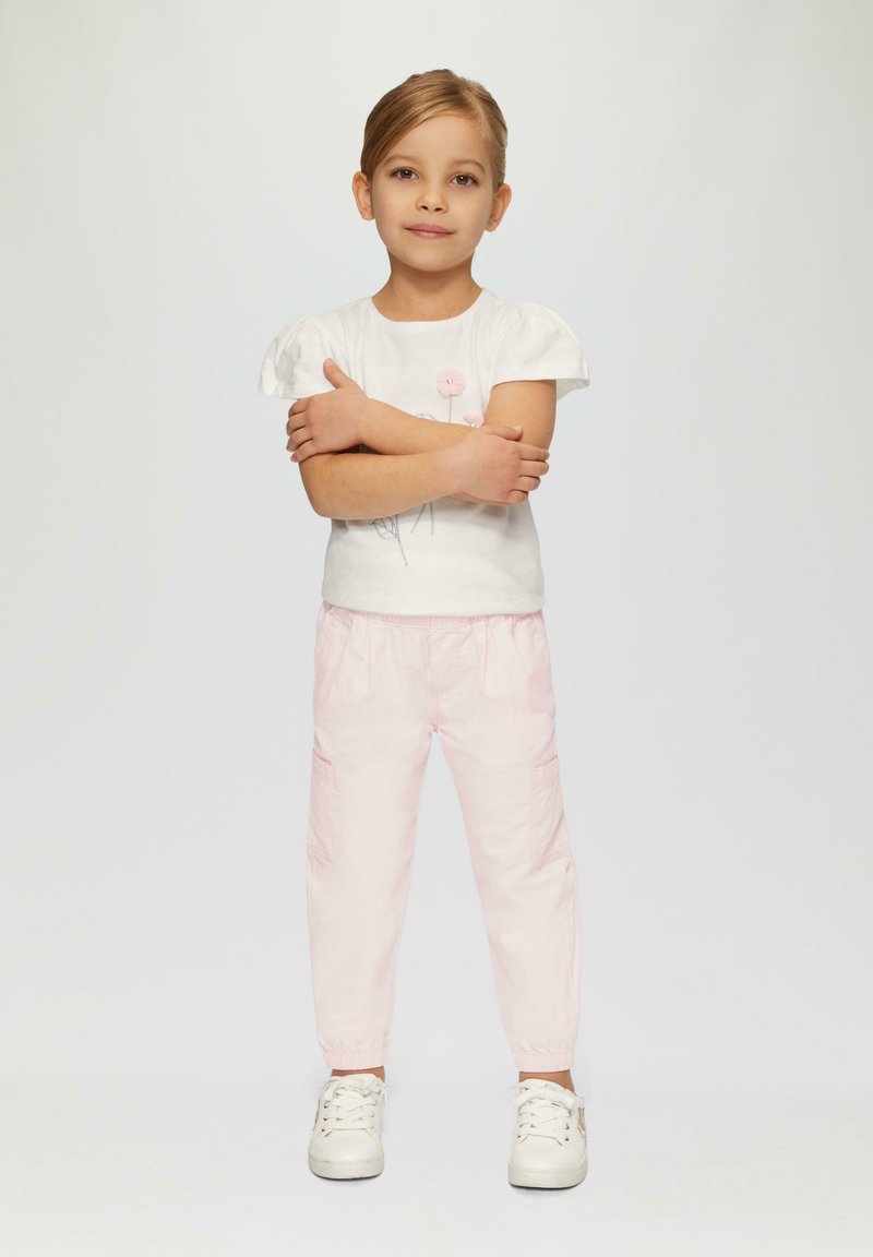 T-shirt blanc à manches courtes avec un design floral, associé à un pantalon cargo rose clair à taille élastique. Modèle portant des baskets blanches, les bras croisés.