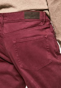 Pantalons en coton bordeaux avec une texture lisse, dotés d'une étiquette en cuir à la taille et d'une seule poche arrière avec un contour brodé.