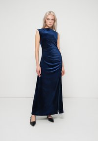 Γυναίκα στέκεται φορώντας ένα αμάνικο, μακρύ βελούδινο φόρεμα σε σκούρο ναυτικό χρώμα με ρυάκι λεπτομέρειες, σε συνδυασμό με μαύρες γόβες με μυτερή μύτη, σε λευκό φόντο.