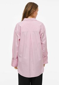 Šviesiai rožinė marškinė su ilgomis rankovėmis, nugaros klostėmis ir užsegamomis rankovėmis, su klasikiniu apykakle ir šiek tiek išlenktu sijonu.