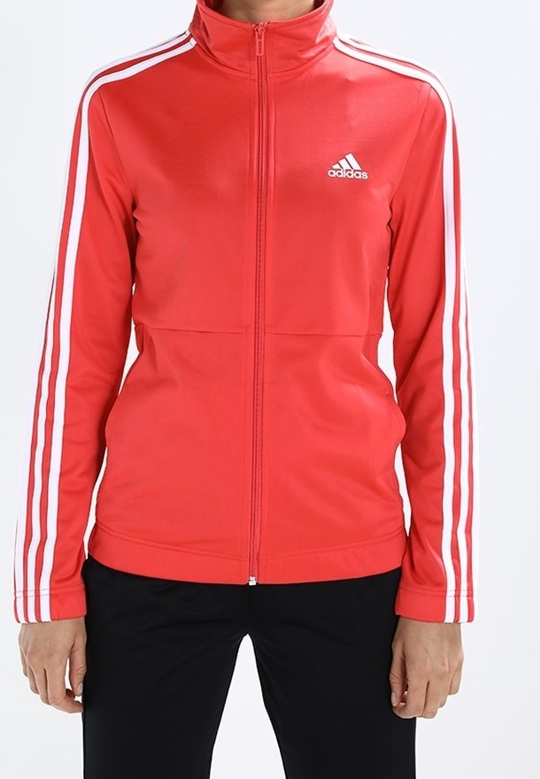 Veste zippée rouge Adidas avec des bandes blanches sur les manches et un logo sur la poitrine, portée avec un pantalon noir.