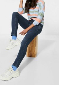 Tajta jeans i mörkblå denim, bärs med en flerfärgad randig stickad tröja och vita sneakers. Sitter på en träpall.