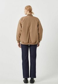 Chaqueta bomber marrón con puños y dobladillo elásticos, mostrada desde la parte trasera. Combinada con jeans rectos de color azul oscuro y botas negras hasta el tobillo.
