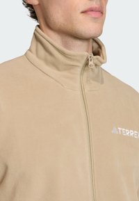 Beige fleece jack met een hoge kraag, ritssluiting aan de voorkant en het 'TERREX' logo in het wit. Zachte textuur zonder zichtbare patronen of accenten.