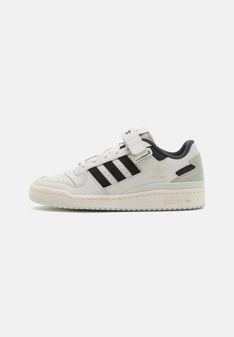 Låg Adidas-sneaker i off-white med svarta ränder, snörning fram och kardborreband, visad i högersidoprofil mot en enfärgad bakgrund.