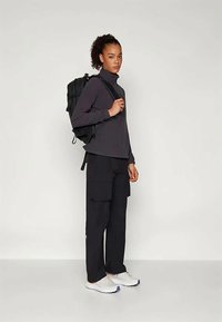 Jauna moteris stovi, vilkinti juodus cargo kelnes, tamsią ilgaaunę maikę, baltus sportinius batelius ir nešanti juodą kuprinę per vieną petį.