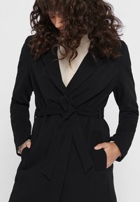 Černý kabát s páskem, klopovým límečkem, dvěma předními kapsami a texturovanou tkaninou. Obsahuje detail uzlu v pase.