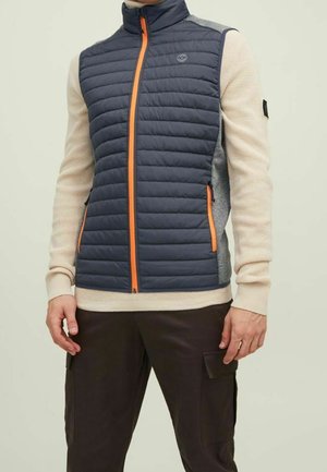 Man draagt een marineblauwe doorgestikte bodywarmer met oranje rits en zakken over een beige trui en donkere cargobroeken, staand met ontspannen handen.