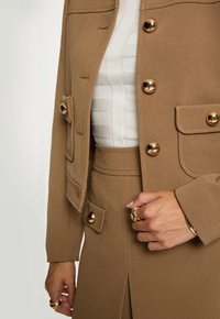Vrouw in een camel-kleurig pakken met gouden knopen, gecombineerd met een witte textuurtop en gouden ringen op beide handen.