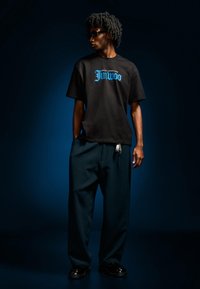 Černé tričko s krátkým rukávem s modrým potiskem, doplněné o zelené široké kalhoty a černé boty s malým řetízkovým detailem.