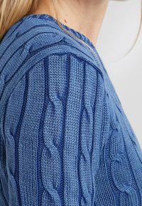 Närbild av en person som bär en blå, tjock kabelstickad tröja med flätade och snurrade sömmönster.