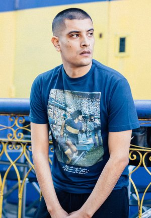 Marineblauw t-shirt met een grote afbeelding van een voetballer te midden van een juichende menigte, met daaronder tekst in een contrasterende kleur.