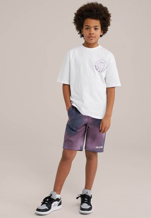 T-shirt bianco in cotone con grafica viola e design loose-fit, abbinato a pantaloni corti con gradiente viola caratterizzati da un motivo a punti e logo bianco.