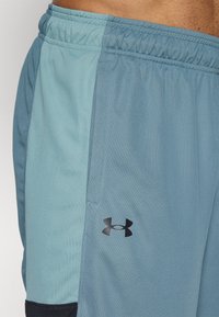 Κοντινό πλάνο ενός ατόμου που φοράει αθλητικά σορτς δίχρωμα σε μπλε αποχρώσεις με ελαστική μέση και το λογότυπο Under Armour στην αριστερή πλευρά.