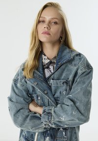 Blonde vrouw met los haar, draagt een oversized spijkerjasje met verfspetters en een gestreept overhemd met kraag, met gekruiste armen en een neutrale uitdrukking.