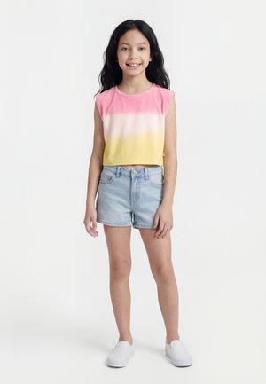 Dziewczyna stoi i uśmiecha się, ma na sobie różowo-biało-żółtą bluzkę bez rękawów, jasne szorty z denimu oraz białe buty wsuwane na stopę na białym tle.