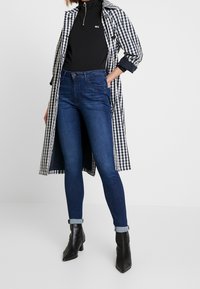 Jean skinny taille haute, bleu foncé, associés à un haut noir zippé et un long manteau à carreaux, rehaussés par des bottes à talons noires.