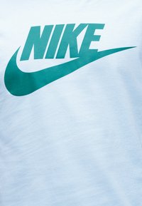 Λογότυπο της Nike με έντονο τιρκουάζ κείμενο και σύμβολο swoosh σε φόντο από ανοιχτό μπλε ύφασμα.