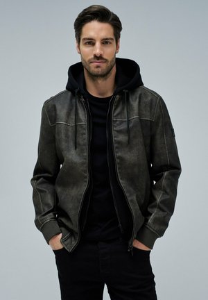 Hombre con barba y cabello oscuro, vistiendo una sudadera con capucha negra y chaqueta gris oscuro con cremallera, de pie con las manos en los bolsillos contra un fondo liso.