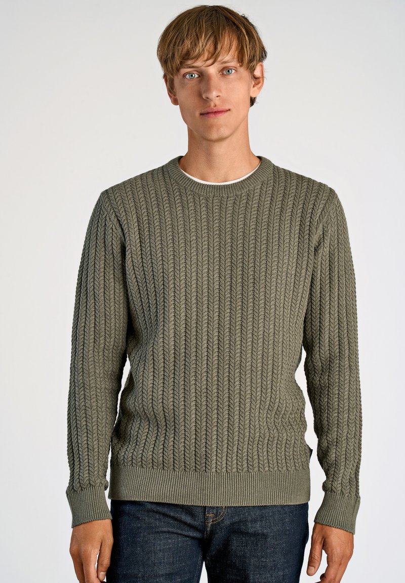 Pull en laine verte tricoté avec un design en câble texturé, col rond et poignets et ourlet côtelés, assorti à un jean en denim foncé.