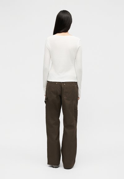 Γυναίκα που στέκεται με στραμμένη την πλάτη, φορώντας λευκή μπλούζα με μακριά μανίκια και καφέ φαρδιά παντελόνια με πολλές τσέπες.