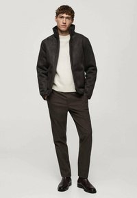 Dunkelbraune Zip-Jacke mit hohem Kragen, getragen über einem cremfarbenen Pullover und braunen karierten Hosen; das Model steht mit den Händen in den Taschen.