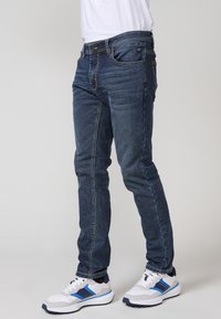 Jeans de mezclilla azul oscuro con corte ajustado, cinco bolsillos, costuras doradas y cremallera. Llevados con zapatillas blancas con detalles azules.