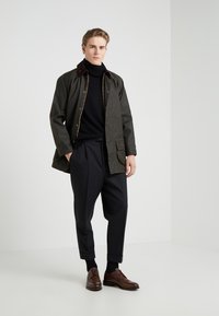 Dark green, waterproof jacket with brown accents, zip and snap closure, paired with a black turtleneck and tailored black pants. Brown leather shoes.
