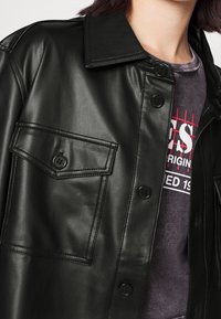 Černá kožená bunda s špičatým límcem, předními knoflíky a jednou kapsou na prsu. Hladký povrch s viditelnými švy.