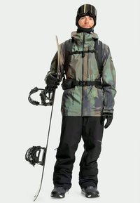 Mann in Camouflagejacke, schwarzen Hosen, Handschuhen und Brille hält ein Snowboard in aufrechter Position vor einem einfarbigen Hintergrund.