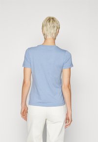 T-shirt azzurro chiaro a maniche corte con scollatura rotonda, in tessuto morbido e dal design aderente, visto da dietro. Pantaloni bianchi visibili.