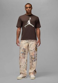 Camiseta de algodón marrón con el logo blanco de Jordan, combinada con pantalones crema que presentan patrones de graffiti coloridos. Calzado: zapatillas blancas.