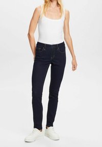 Žena v tmavě modrých skinny jeans, bílém bezramínkovém topu a bílých teniskách stojí proti jednoduše pozadí.