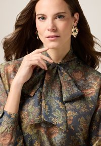 Blommig blus i mörkblått med ett stort rosettband vid halsen. Har jordnära toner av grönt, orange och brunt. Modellen bär dekorativa örhängen.