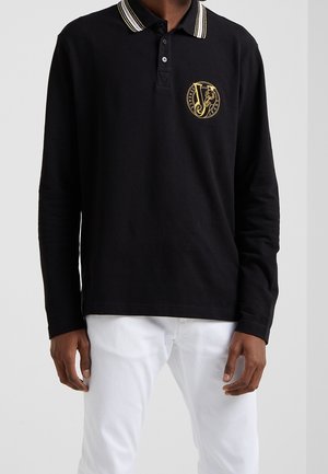 Polo noir à manches longues avec un col rayé, présentant un logo brodé sur la poitrine. Porté avec un pantalon blanc pour un contraste.