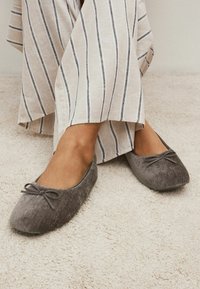 Grå sammet balletflade med en rund tå og en lille sløjfeaccent foran. Båret med stribet beige linned, præsenteret på en struktureret overflade.