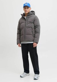 Młody mężczyzna w szarej puchowej kurtce, czarnych spodniach, białych trampkach i niebieskiej czapce z daszkiem stojący na tle jednolitego jasnego tła.