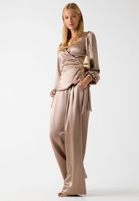 Frau trägt glänzende beige Satin-Wickelbluse und weite Hosen, hält kleine metallisch goldene Schultertasche, steht vor weißem Hintergrund.