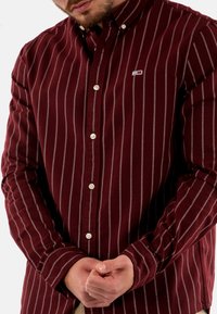 Camicia button-up bordeaux con strisce verticali grigie, dotata di colletto aperto e bottoni bianchi. Include un piccolo logo sul lato sinistro del petto.